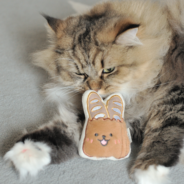 A fluffy long-haired cat lies on a soft gray surface, chewing on a small plush bunny toy with a bread-brown face and embroidered details.