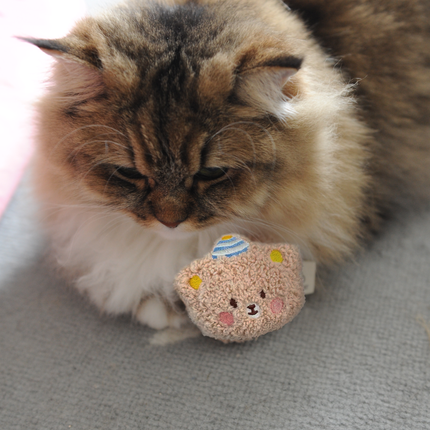 A fluffy long-haired cat with brown, black, and cream fur is lying on a gray textured surface and gently holding a small crocheted teddy bear toy.