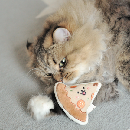 A fluffy long-haired cat lies on a light gray carpet, chewing on a plush toy shaped like a slice of pizza with a cat face.