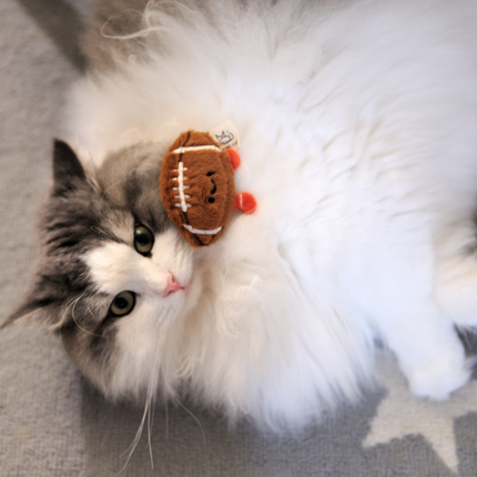 A fluffy long-haired cat with gray and white fur lies on a gray patterned rug, holding a small brown felt football toy near its face.