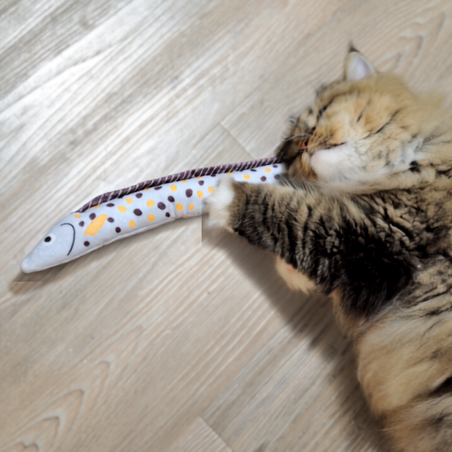 A fluffy cat lies on a light wood floor, holding and biting a long, fish-shaped plush toy with a white body, dark spots, and yellow accents.