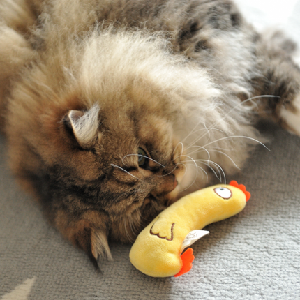 A fluffy long-haired cat lying on a gray carpet next to a small yellow plush toy with orange accents.
