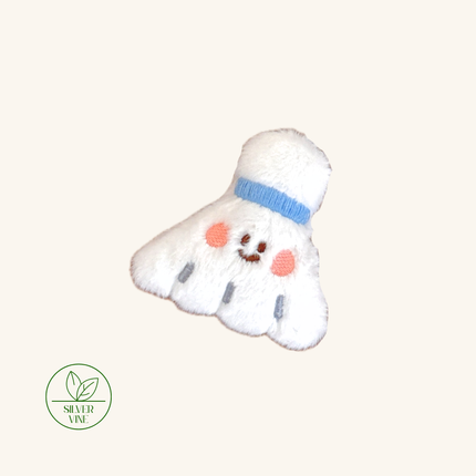 A plush embroidered shuttlecock-shaped accessory in white with a light blue band, pink embroidered cheeks, and a small stitched smile. A green 'Silver Vine' logo appears in the lower-left corner on a plain warm off-white background.
