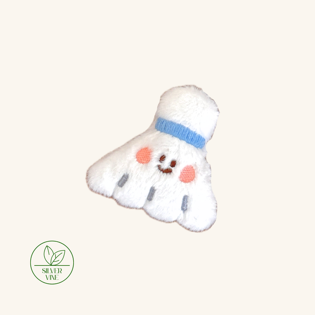 A plush embroidered shuttlecock-shaped accessory in white with a light blue band, pink embroidered cheeks, and a small stitched smile. A green 'Silver Vine' logo appears in the lower-left corner on a plain warm off-white background.