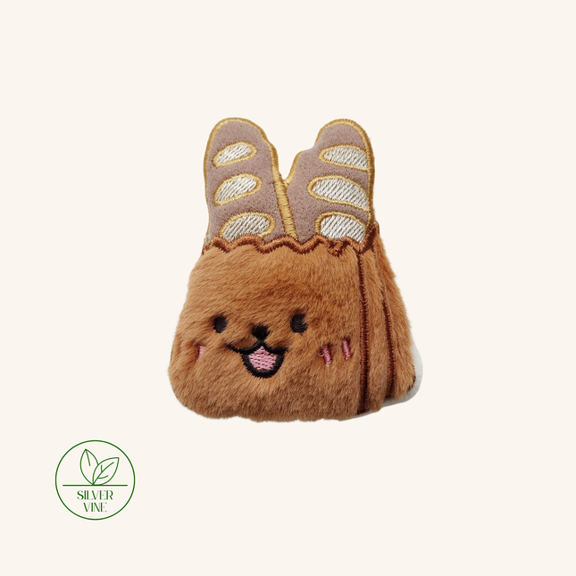 A small plush bunny-shaped accessory in warm brown faux fur, styled like a character with embroidered facial features and a top section resembling beige and white striped bread or pastries. The item is centered on a plain off-white background with a green 'Silver Vine' logo watermark in the bottom-left corner.