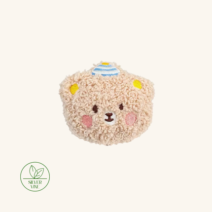 A plush teddy bear head coin purse in a textured beige sherpa fabric, featuring embroidered details of a smiling face, yellow ears, pink cheeks, and a blue and yellow striped hat. A green 'Silver Vine' brand logo is visible in the bottom-left corner against a solid cream background.