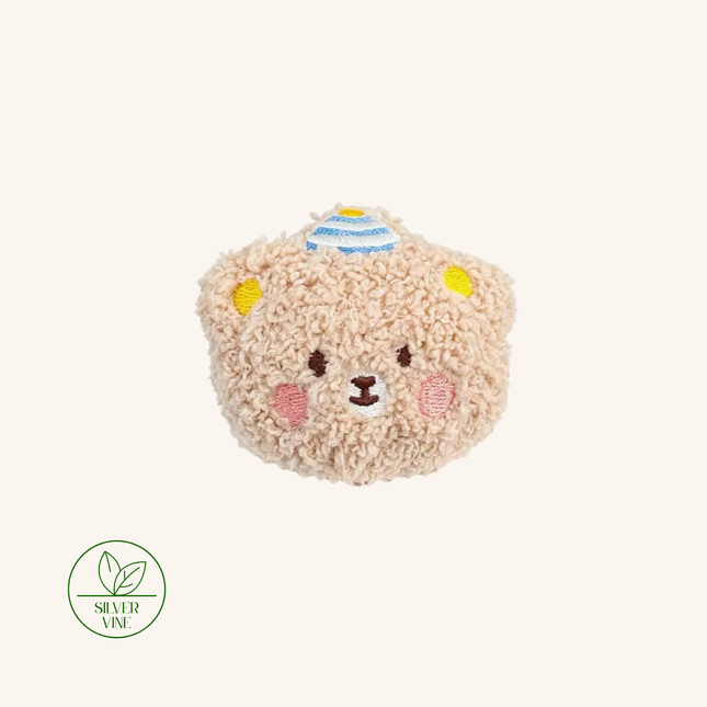 A plush teddy bear head coin purse in a textured beige sherpa fabric, featuring embroidered details of a smiling face, yellow ears, pink cheeks, and a blue and yellow striped hat. A green 'Silver Vine' brand logo is visible in the bottom-left corner against a solid cream background.