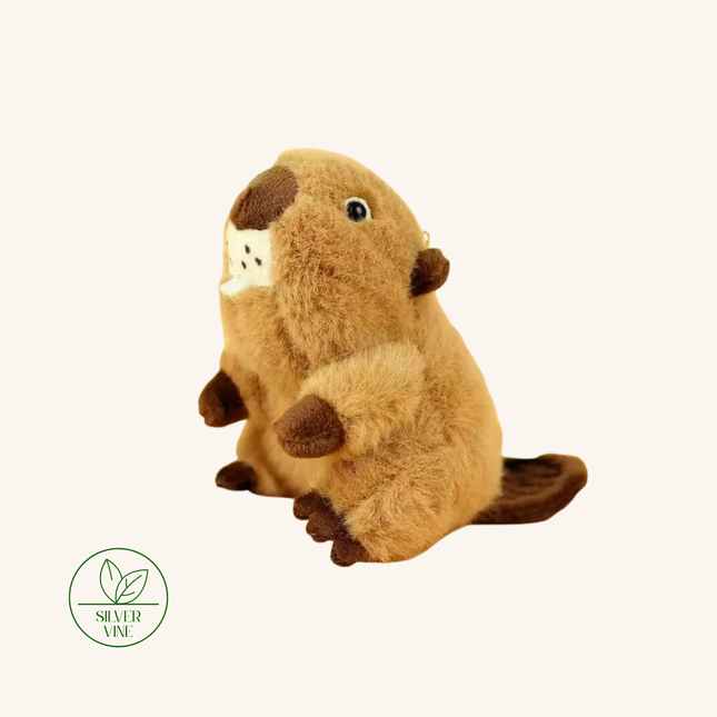 A plush toy beaver in a sitting position, made of soft brown faux fur with darker brown paws, snout, and tail, and a small white patch on its chest. The beaver is photographed on a solid off-white background with a 'Silver Vine' watermark in the bottom-left corner.