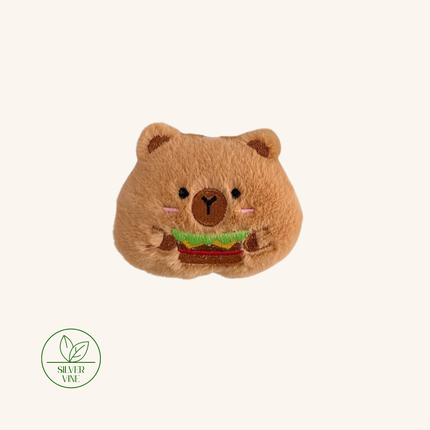 A plush bear-shaped hair accessory in a warm brown color, holding a small embroidered burger. The bear has embroidered black eyes, a brown nose, and pink cheeks.