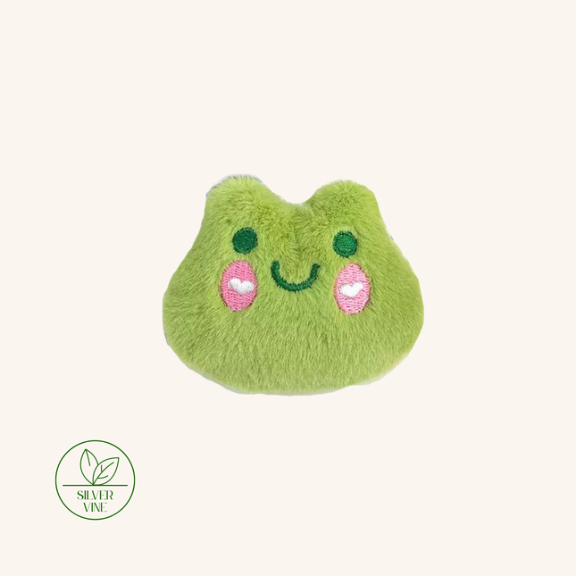 A plush, fuzzy green pouch or accessory shaped like a smiling cartoon face, shown on a plain light beige background. The face has embroidered dark green eyes and mouth, and two pink heart-shaped cheeks.