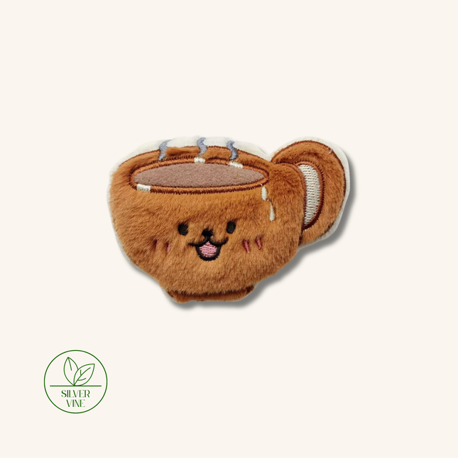 A plush brown cup-shaped accessory with embroidered facial features, small silver metallic details, and a white handle, shown on a plain light beige background. A green 'Silver Vine' logo appears in the lower-left corner.